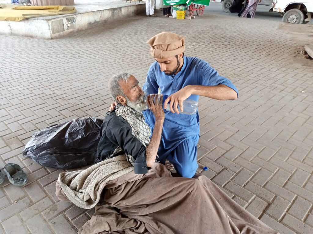 A young man helps a homeless senior by giving water outdoors in Karachi, Pakistan.