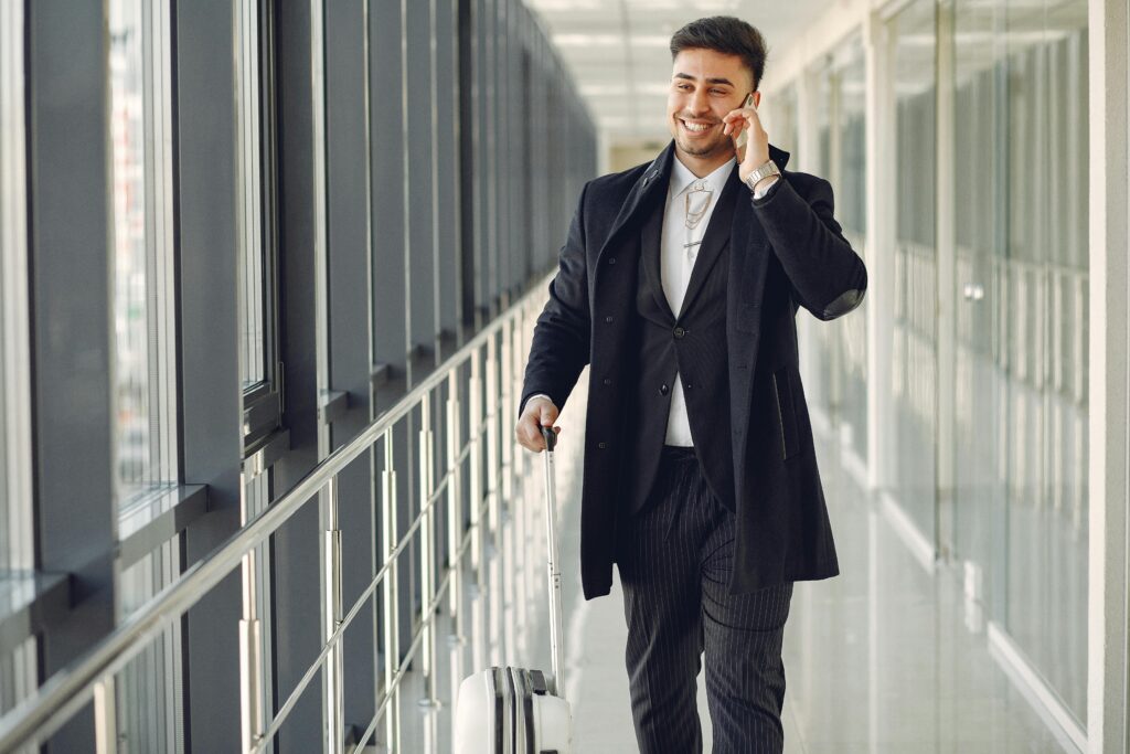 Happy ethnic male in formal suit standing in modern hallway while holding suitcase in hand and talking on mobile phone